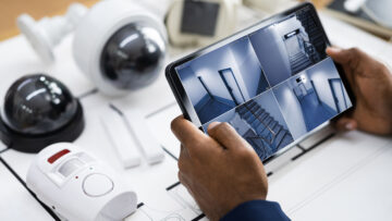 A close up shot of someone holding a tablet showing CCTV footage. There are security cameras in the background