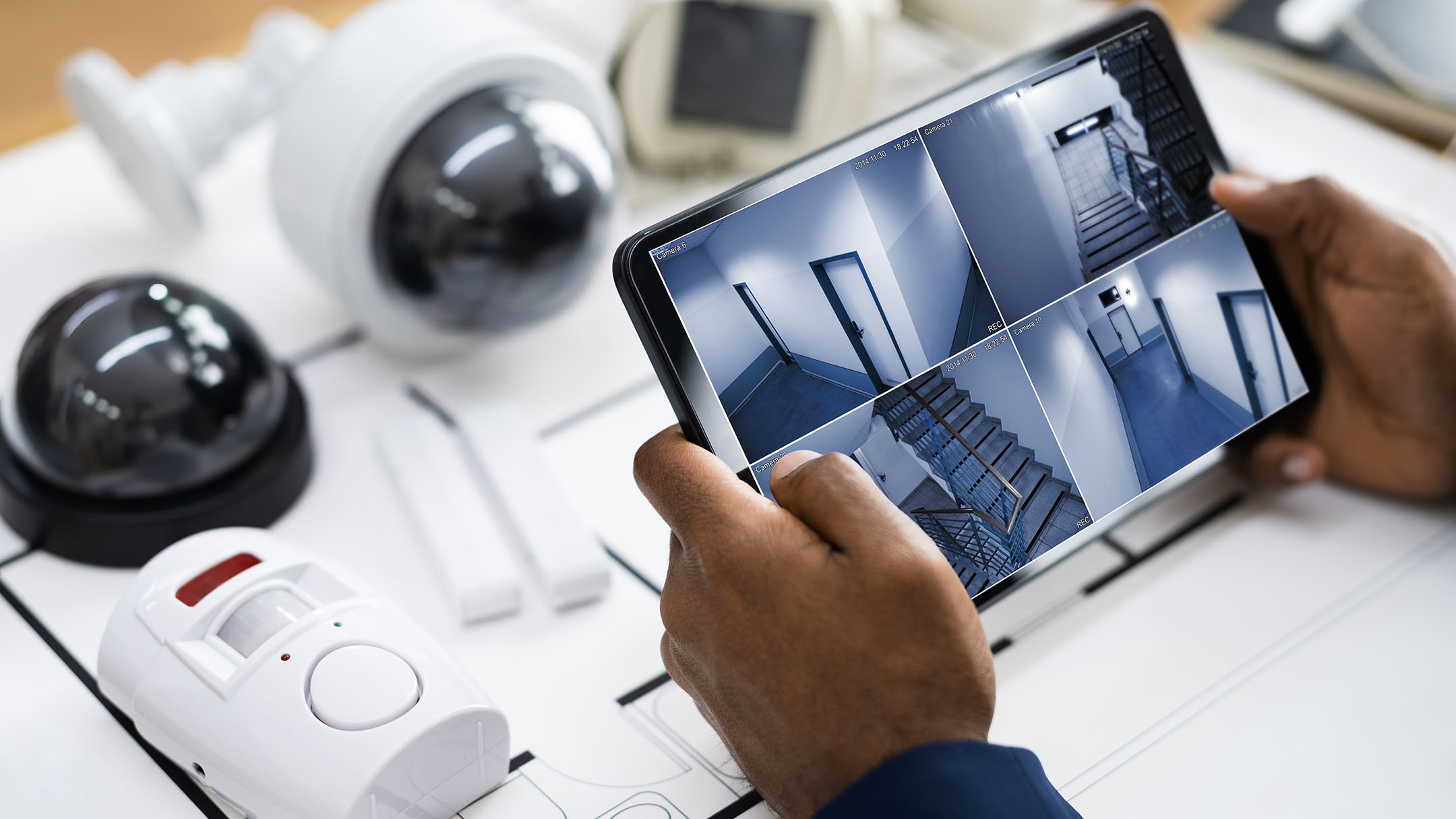 A close up shot of someone holding a tablet showing CCTV footage. There are security cameras in the background